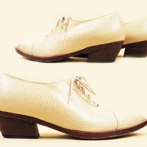 May include: A pair of cream-colored leather shoes with a lace-up front and a low block heel. The shoes have a textured, snakeskin-like pattern.