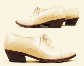 May include: A pair of cream-colored leather shoes with a lace-up front and a low block heel. The shoes have a textured, snakeskin-like pattern.