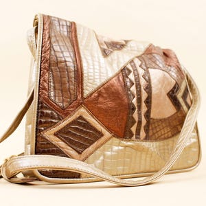 May include: A shoulder bag with a patchwork design. The bag features various shades of brown, tan, and cream leather, with a textured, crocodile-like pattern on some panels. The bag has a long, gold-colored strap.