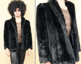 May include: A black faux fur jacket with a shawl collar. The jacket is unbuttoned and has a black mesh shirt underneath.