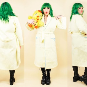 May include: A pale yellow quilted robe with light blue trim and pockets. The robe has a matching belt and is knee-length. The model is wearing black boots and holding a yellow stuffed animal.