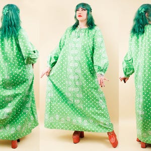 May include: A floor-length, long-sleeved dress in bright green with a white bandana-style print featuring paisley motifs and polka dots. The garment has a loose, flowing silhouette, a gathered neckline with a tie closure, and cuffed sleeves.