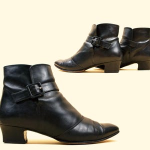 May include: A pair of black leather ankle boots with a buckle strap. The boots have a low heel and a pointed toe.