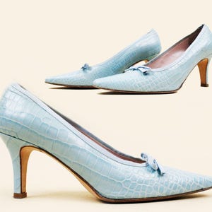 May include: A pair of light blue, crocodile-embossed pumps with a pointed toe and a small bow detail on the vamp. The heels are approximately 3 inches high.
