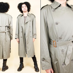 May include: A light green trench coat with a double-breasted design, featuring a belt, epaulets, and button details. The coat has a classic silhouette and is made of a smooth fabric. The coat is approximately 55 inches long.