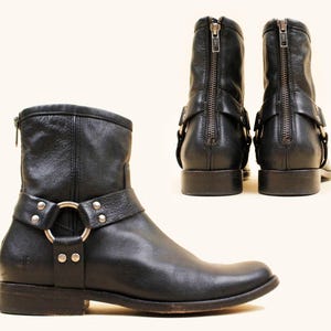 May include: Black leather ankle boots with a harness strap and silver-tone hardware. The boots have a back zipper and a low heel. One boot is shown from the side, and two boots are shown from the back.