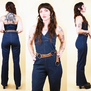 May include: A dark blue denim jumpsuit with a halter neck and flared legs. The jumpsuit has a brown leather belt with silver studs and a silver buckle. The model is wearing the jumpsuit in a studio setting.