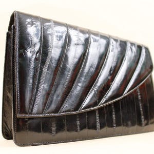 May include: A black clutch with a glossy finish. The clutch features a flap closure and a textured, ribbed design across the front. The bag is rectangular and appears to be made of a shiny material.