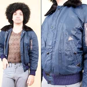 May include: A navy blue bomber jacket with a black faux fur collar, featuring a zipper pocket on the left sleeve and a purple ribbed hem and cuffs. The jacket has a button-up front and a triangular patch detail.