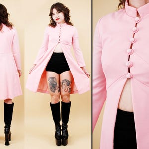 May include: A light pink long-sleeved coat dress featuring a high mandarin collar and a front button closure with five round matching buttons. The garment has a fitted bodice that transitions into a flared skirt, with a split opening at the front.