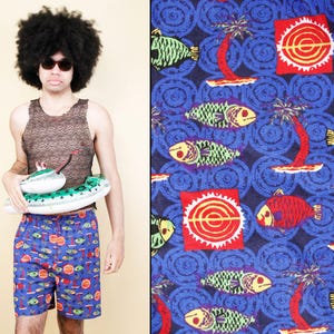May include: A man wearing a brown tank top and blue swim trunks with a fish and palm tree pattern. He is holding a green and white inflatable pool float shaped like a submarine. The background is a blue and white patterned fabric with fish and palm trees.