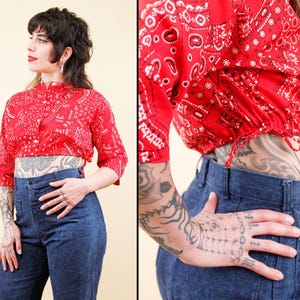 Puede incluir: Un top corto rojo con estampado de bandana, cuello mandarín y cierre de botones. El top tiene mangas tres cuartos. Combinado con pantalones vaqueros de cintura alta y azul oscuro.