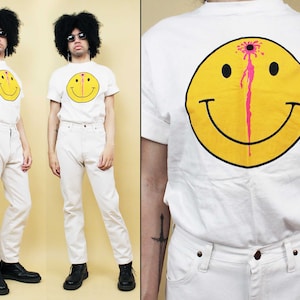 May include: White t-shirt with a yellow smiley face graphic with a pink blood splatter on the face. The smiley face has a black dot for each eye and a black line for the mouth.