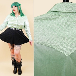 May include: A long-sleeved, light mint green button-down shirt featuring a pointed collar and a textured, woven V-shaped yoke panel across the chest and shoulders. The shirt is paired with a plain black skater skirt and a black leather belt.