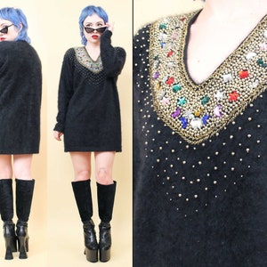 May include: A black fuzzy sweater dress with a V-neck and a gold and jewel-toned beaded embellishment around the neckline. The model is wearing black platform boots.