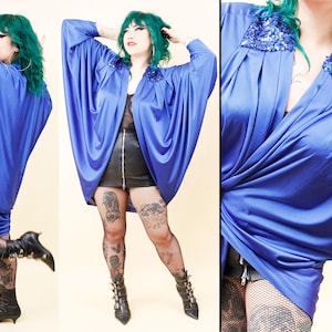 May include: A striking blue draped top with sequin detailing on the shoulders. The top features wide, flowing sleeves and a deep V-neck. The model is wearing black boots and fishnet stockings. The outfit is a bold fashion statement.