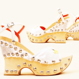 May include: A pair of platform sandals with wooden soles and white fur accents. The sandals feature silver studs along the edges and white straps with buckles. The interior is red. The shoes are a unique fashion statement.