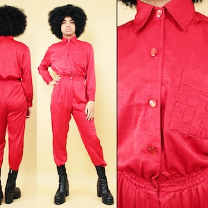 Op de afbeelding: Een felrode jumpsuit met een kraag, lange mouwen en een geweven zakdetail. De jumpsuit heeft een knoopsluiting aan de voorkant en een elastische tailleband. Het model draagt zwarte plateau laarzen. De outfit is een kledingstuk uit één stuk.
