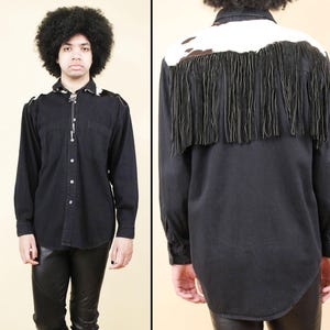May include: Black denim shirt with long sleeves, featuring a white and brown cowhide yoke and long fringe detail across the back. The shirt has two chest pockets and a button-down front. A bolo tie is visible at the collar.