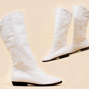 May include: A pair of white knee-high boots featuring a slouchy shaft design, a rounded toe, and a flat black sole. The boots are constructed with a smooth white upper material that extends to the calf, providing a clean and simple aesthetic.