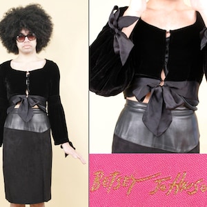 May include: A black velvet long-sleeve top with a square neckline, front tie closure, and ribbon accents on the cuffs, paired with a black skirt featuring a smooth waistband. The garment includes a pink label with the gold text Betsey Johnson.