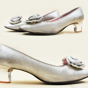 May include: A pair of silver glitter pumps with a clear acrylic heel and a large silver flower embellishment on the toe.
