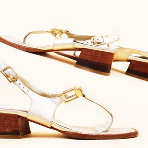 May include: Two pairs of gold-colored sandals with brown wooden block heels. The sandals have a T-strap design with a buckle closure and a clear strap. The heels are approximately 2 inches high.