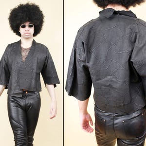 Può includere: Giacca nera in stile patchwork con scollo a V e maniche corte. La giacca ha un aspetto strutturato ed è abbinata a pantaloni di pelle neri. L'outfit è indossato da una persona con afro e occhiali da sole.
