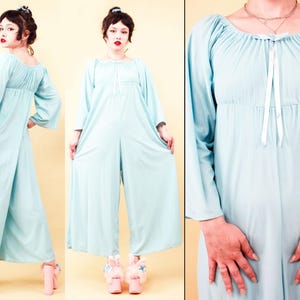 May include: A light blue jumpsuit with long sleeves and wide legs. The top features a gathered neckline with a white ribbon tie. The model is wearing pink platform shoes. The jumpsuit is made of a soft, flowing fabric.