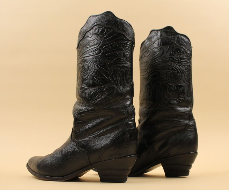May include: A pair of black leather cowboy boots with intricate embossed designs. The boots have a pointed toe and a low heel.