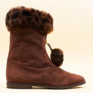 May include: Brown suede boots with a faux fur cuff featuring a leopard print pattern. The boots have a zipper on the side and a fluffy pom-pom detail.