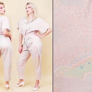 May include: A light pink jumpsuit with short sleeves and a button-down top. The pants are high-waisted and tapered. The fabric has a subtle pattern and a matching belt. The outfit is paired with white heels.