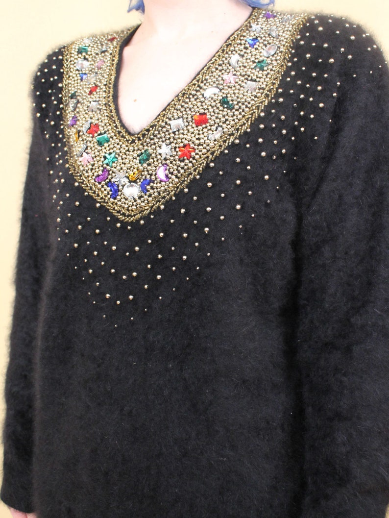 May include: A black fuzzy sweater with a v-neck and a decorative beaded neckline featuring colorful rhinestones and gold accents.