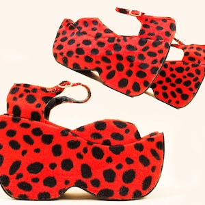 May include: A pair of platform sandals with a bold red and black spotted pattern. The sandals have a thick sole and an ankle strap with a gold buckle. The open-toe design and unique pattern make these shoes a statement piece.