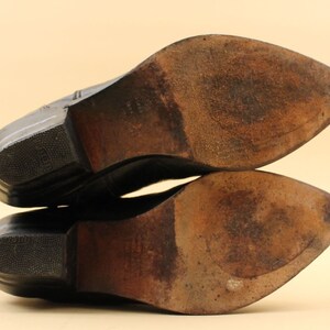 May include: Close-up of the soles of a pair of black leather cowboy boots. The boots have a pointed toe and a stacked heel.