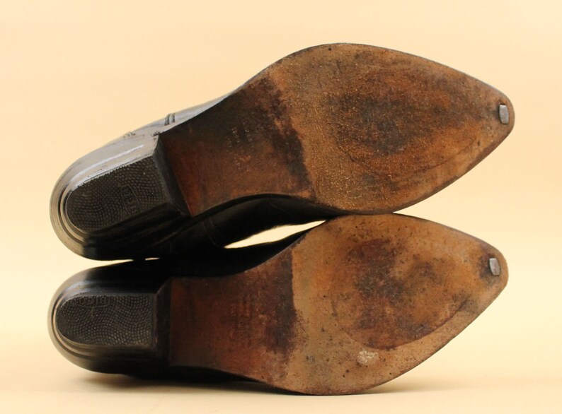 May include: Close-up of the soles of a pair of black leather cowboy boots. The boots have a pointed toe and a stacked heel.