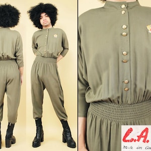 Op de afbeelding: Een groene jumpsuit met knopen en een geplooide taille. De jumpsuit heeft lange mouwen en een hoge hals. Het model draagt zwarte laarzen.