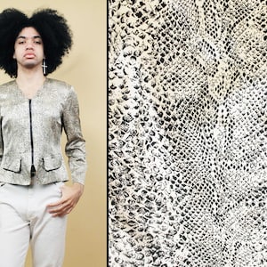 Puede incluir: Chaqueta dorada y negra con estampado de piel de serpiente, con cremallera y ribete negros. La chaqueta tiene cuello en V y dos bolsillos pequeños. La chaqueta se combina con pantalones blancos. La imagen también muestra un primer plano del estampado de piel de serpiente.