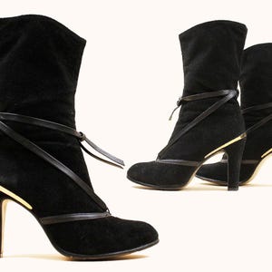 May include: Black suede ankle boots with a high heel and a leather strap detail. The boots have a gold accent on the heel and a pointed toe. The boots are approximately 4 inches high.