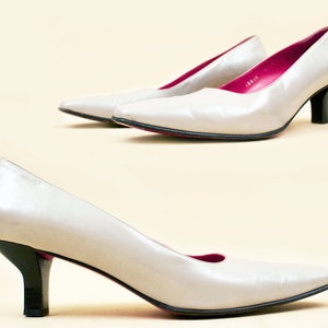 May include: A pair of white leather pumps with a pointed toe and a low heel. The shoes have a pink lining and a black heel.