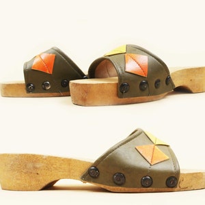 May include: A pair of green leather platform clogs with orange and yellow geometric designs. The clogs have a wooden platform sole and are fastened with metal studs.