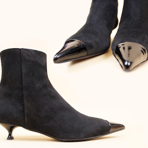 May include: Black ankle boots with a pointed toe and a glossy black cap. The boots have a low, curved heel and are made of a suede-like material. The image shows the boots from multiple angles.
