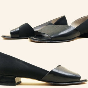 Puede incluir: Un par de sandalias de tacón de cuero negro con puntera abierta y una tira de tela negra en la parte superior. Las sandalias tienen un tacón de bloque bajo.