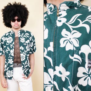 Puede incluir: Una camisa hawaiana verde y blanca con cierre de cremallera. La camisa tiene un estampado floral con flores blancas sobre un fondo verde. La camisa tiene cuello y mangas cortas.
