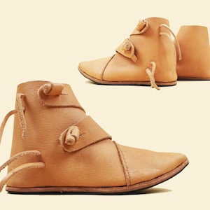 Può includere: Un paio di stivaletti in pelle marrone chiaro con punta. Gli stivali presentano cinturini e lacci decorativi, con un design semplice. Le calzature sono probabilmente fatte a mano, con cuciture visibili e un'estetica naturale e terrosa.