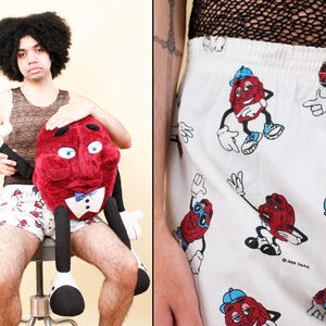 May include: A person holding a large plush raisin character with a blue bow tie and white gloves. The person is wearing a patterned tank top and white shorts with a raisin cartoon print.