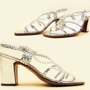 May include: A pair of silver high-heeled sandals with a woven strap design. The sandals have a chunky heel and an open toe.
