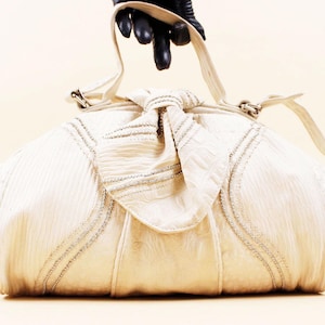 May include: Cream-colored handbag with a textured, gathered design. The bag features silver-toned embellishments and a knotted closure. A long, thin strap extends from the top. The bag is held by a gloved hand.