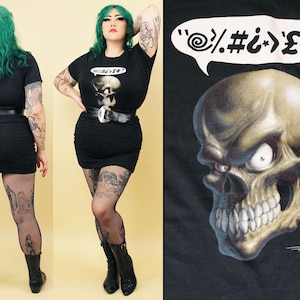 May include: Black t-shirt with a skull graphic and the text "'/.#;>£#!". The skull has a menacing expression and is surrounded by a speech bubble with the text "'/.#;>£#!".