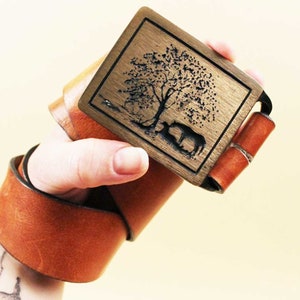 May include: A brown leather belt with a brass buckle featuring a laser-engraved design of a tree and two animals.